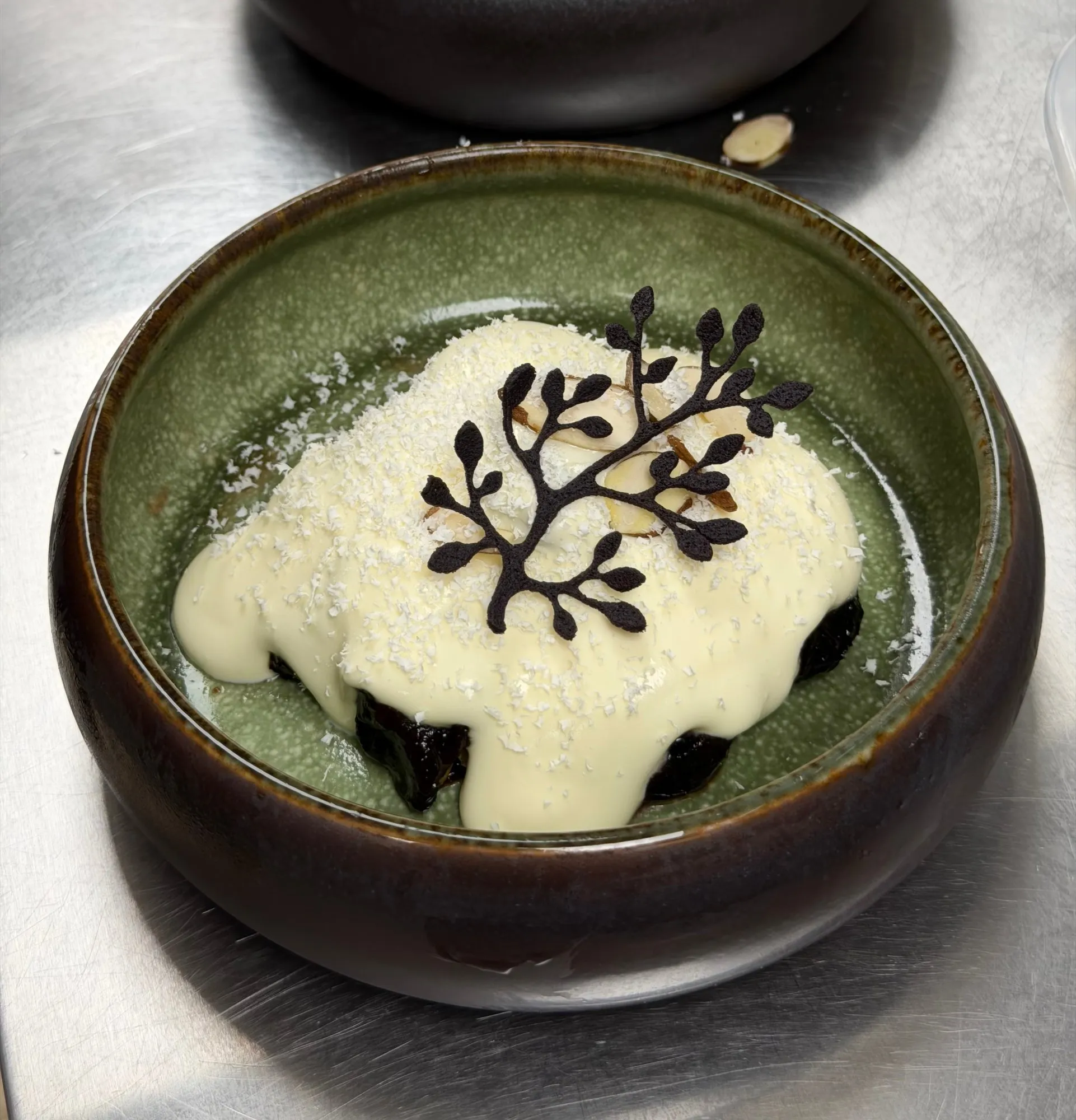 Prunes under white chocolate sauce in a green ceramic bowl, with a dark chocolate branch on top